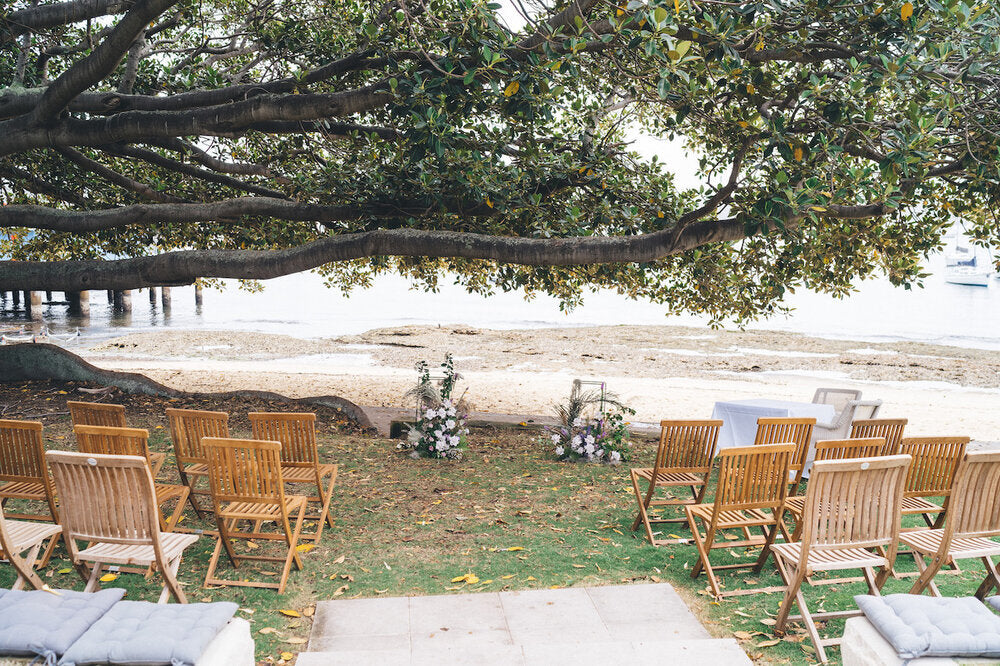 Ceremony set up under the Moreton Bay fig tree at Robertson Park, Watsons Bay, with floral clusters and timber chairs