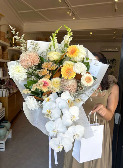 Bouquet of muted, cream, buttery, nude fresh flowers wrapped in white paper, ready for delivery, with a white gift bag held by a person in a store setting.