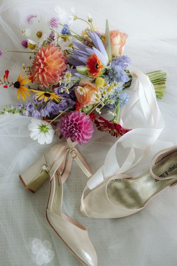 summery bright colourful garden inspired bridal bouquet flat lay with bridal wedding shoes and trailing satin ribbons