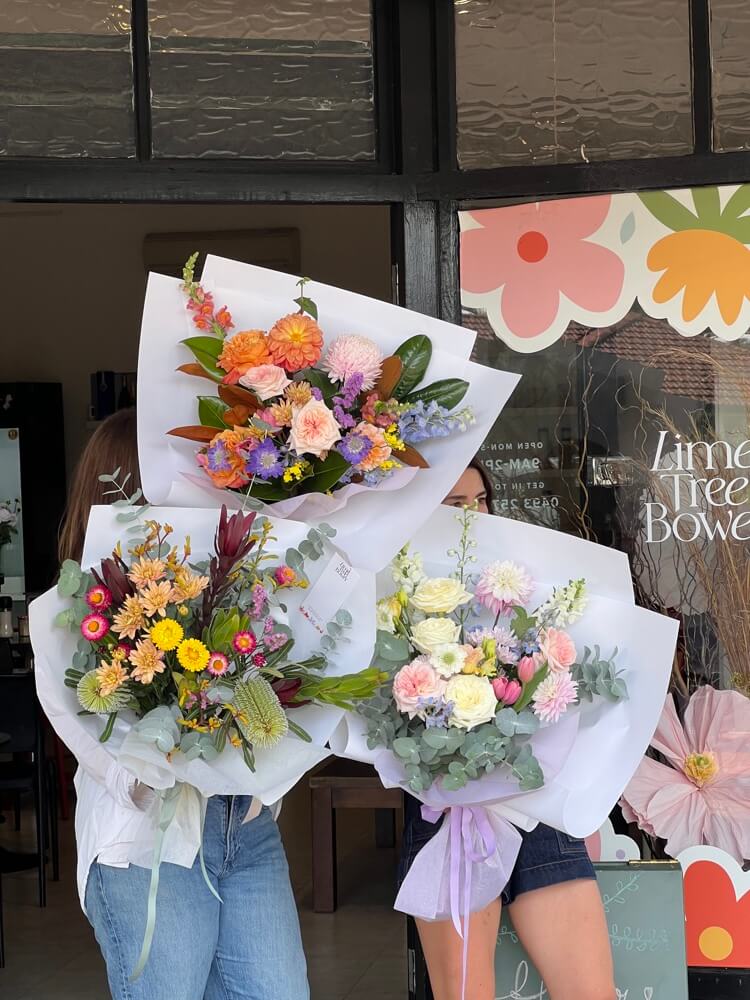 Two people holding colourful flower bouquets for Mothers Day in front of a store window with 'Lime Tree Bower Florist' flower shop branding.